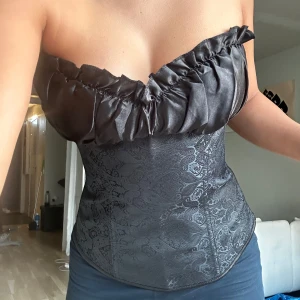 Svart korsett med spetsdetaljer - Snygg svart korsett med spetsdetaljer och volang upptill. Den har en tight passform och snörning i ryggen för justerbar storlek. Aldrig använd då den var menad till halloween men kom försent + är för stor vid brösten för mig. 