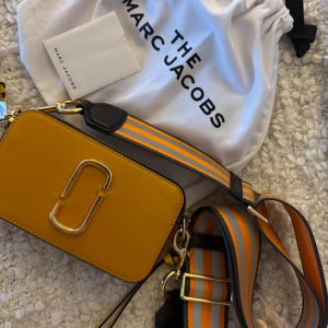Gul axelväska från Marc Jacobs - Säljer en superfin gul axelväska från Marc Jacobs. Den har en cool randig axelrem i orange och blått, och snygga gulddetaljer. Perfekt för att liva upp vilken outfit som helst! Väskan är i nytt skick och kommer med en dustbag.