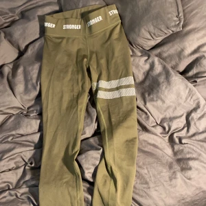 Gröna leggings från Stronger - Säljer ett par gröna leggings från Stronger i storlek XS. De är tillverkade av 75% återvunnen polyester och 25% elastan, vilket gör dem superstretchiga och bekväma. De har ett coolt prickigt mönster på ena benet och en bred resår i midjan med Stronger-loggan. Perfekta för träning eller en chill dag hemma!