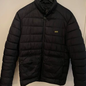 Svart quiltad jacka från Barbour - Säljer en snygg svart quiltad jacka från Barbour International. Den har en dragkedja framtill och två praktiska fickor med knappar. Perfekt för höst och vår, och den är i mycket bra skick.