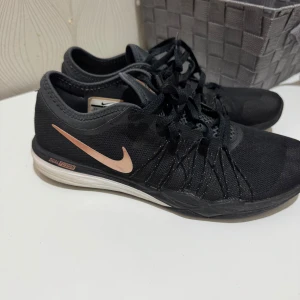 Svarta Nike träningsskor - Säljer ett par svarta Nike träningsskor i bra skick. De har en snygg design med svart mesh och en diskret rosa Nike-logga på sidan. Skorna har snörning och en bekväm sula som passar perfekt för träning. Perfekta för gymmet eller löprundan!