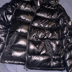 Svart pufferjacka från Moncler - Säljer en snygg svart pufferjacka från Moncler i mycket bra skick. Jackan har en glansig finish och är perfekt för kalla vinterdagar. Den har en dragkedja framtill och praktiska fickor med dragkedjor. Huvan är justerbar för extra skydd mot väder och vind. Perfekt för både vardag och fest!