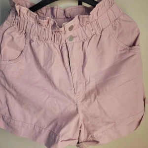 Ljusa rosa shorts från H&M - Säljer ett par ljusrosa shorts från H&M i storlek 40. De har en högmidjad design med elastisk midja och två knappar framtill. Perfekta för sommardagar med en avslappnad och bekväm passform. Materialet känns mjukt och luftigt, idealiskt för varma väder.