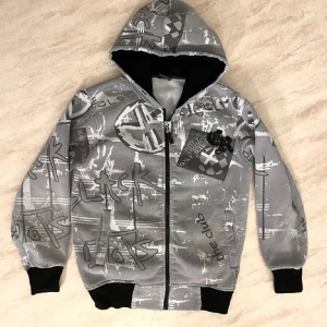 Grå mönstrad zipup hoodie  - Säljer en grå hoodie med coolt grafiskt mönster och textdetaljer. Den har en dragkedja framtill och en skön huva. Rätt så grisch soho om du frågar mig iaf 