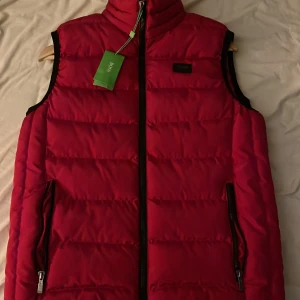 Röd dunväst från Hugo Boss - Säljer en snygg röd dunväst från Hugo Boss i nytt skick. Västen har en dragkedja framtill och två praktiska fickor med dragkedjor. Perfekt för höst och vår när du vill hålla dig varm och stilren. Passar både till vardags och lite finare tillfällen.   PRISET GÅR ATT DISKUTERA!