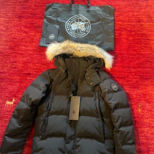 Svart dunjacka från Canada Goose - Säljer en svart dunjacka från Canada Goose i toppskick. Jackan har en fluffig pälsdetalj på huvan och stängs med både dragkedja och knappar. Perfekt för kalla vinterdagar! 🖤