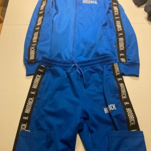 Hoodrich tracksuit - Säljer en blå träningsdräkt från Hoodrich i mycket bra skick. Setet består av en jacka med dragkedja och matchande byxor. Både jackan och byxorna har svarta band med Hoodrich-loggan längs ärmar och ben. Perfekt för en sportig look eller avslappnad stil. Tröja M byxor L