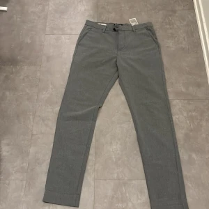 Grå chinos från Jack & Jones - Säljer ett par stilrena grå chinos från Jack & Jones i modellen Slim Marco. Byxorna har en slim passform och är perfekta för både vardag och fest. De är i mycket bra skick och har både knappar och dragkedja framtill. Perfekta för höst och vår!
