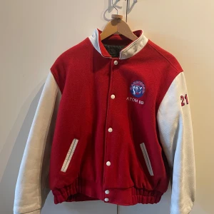 Röd och vit bomberjacka med Lakeshore-märke - Säljer en cool röd och vit bomberjacka med Lakeshore-märke på ryggen och 'ATOM 98' på framsidan. Jackan har vita läderärmar och knappar, samt ribbade muddar. Perfekt för höst och vår! Använt sparsamt, men finns tecken på användning, därav det låga priset. 