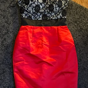 Svart och röd klänning med spetsdetaljer - Snygg klänning med svart spetsöverdel och röd kjol. Perfekt för festliga tillfällen! Den har en tight passform och ärmlös design. Kjolen är i ett glansigt material som ger en lyxig känsla. Storlek XS/S