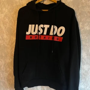 Svart hoodie från FSBN - Säljer en svart hoodie från FSBN i storlek XL. Står 'Just Do Nothing' i vitt och rött på framsidan. Perfekt för en chill dag eller när du bara vill ta det lugnt. Hoodien har en klassisk passform med en stor ficka fram och dragsko i huvan.