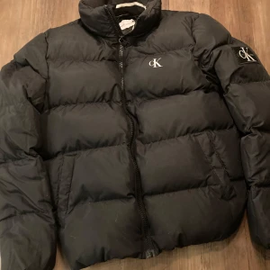 Svart pufferjacka från Calvin Klein Jeans - Säljer en snygg svart dunjacka från Calvin Klein Jeans. Jackan är i mycket bra skick och har en stilren design med CK-logga på bröstet och ärmen. Den är tillverkad av 100% återvunnen polyester och är vattenavvisande. Perfekt för kalla dagar!