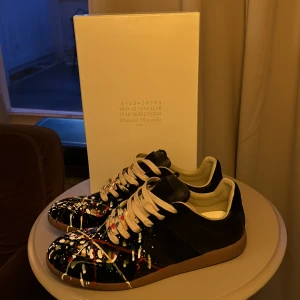 Maison Margiela ”Gats” - Säljer ett par unika Maison Margiela sneakers i svart med coola färgstänk i olika färger. Skorna har en klassisk design med snörning och en bekväm gummisula. Perfekta för att ge din outfit en konstnärlig touch! Kommer med originalförpackning och dustbags. Skicket är 9.5/10, endast använda fåtal gånger! Vid snabb affär så kan jag eventuellt gå ber i pris, tveka inte på att höra av dig vid frågor!❤️🤗 OBS! Endast skorna ingår. Tar gärna emot byten!