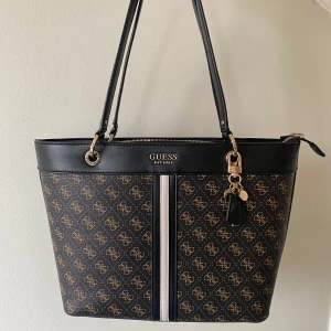 Guess Noelle Elite väska - Guess väska i mycket fint skick, använd ett ytterst fåtal gånger. Den är i fint skick och alldeles för fin att inte användas, tyvärr är det inte min stil längre. Den har ett stort fack i mitten, två stycken mindre fack på insidan samt två små. Den har ett större sidofack och ett litet fack med dragkedja utanpå baktill. Storleken är 40x27x12 cm. 