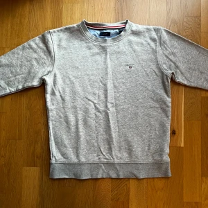 Grå sweatshirt från GANT - Säljer en stilren grå sweatshirt från GANT i mycket bra skick. Tröjan har en klassisk rund halsringning och ett diskret broderat GANT-emblem på bröstet. Perfekt för både vardag och avslappnade tillfällen. Passar bra till jeans eller chinos!