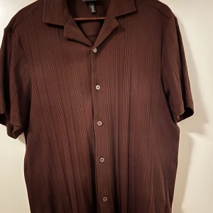 Brun kortärmad skjorta från H&M - Säljer en brun kortärmad skjorta från H&M i regular fit. Skjortan har ett diskret randigt mönster och knappar framtill. Perfekt för både vardag och festliga tillfällen. Materialet känns mjukt och bekvämt mot huden. Använd vid enstaka tillfällen.