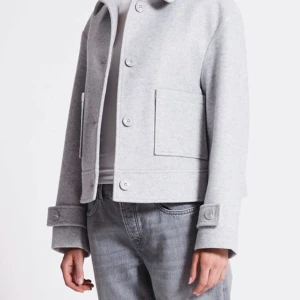 Grå kappa - Säljer en stilren grå kappa i normal passform. Jackan har långa ärmar och stora fickor framtill. Perfekt för höst och vår, med knappar som stängning. Enkelt att matcha med jeans för en casual look.