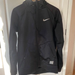 Svart Nike Therma-Fit jacka - Säljer en svart Nike Therma-Fit jacka i mycket bra skick. Den har en dragkedja framtill och två fickor med dragkedja. Perfekt för kyliga dagar med sin värmeisolerande design. Jackan har en huva och är långärmad, vilket gör den idealisk för höst och vår.
