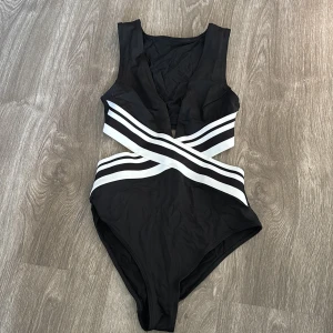 Svart baddräkt med vita band - Snygg svart baddräkt med vita korsade band i midjan. Den har en djup v-ringning och är perfekt för sommarens badäventyr. Materialet känns mjukt och stretchigt för en bekväm passform. Perfekt för både stranden och poolen!