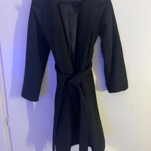 Svart kappa med bälte - Säljer en svart kappa i mycket bra skick, storlek xs från bikbok. Den har ett bälte i midjan för en snygg passform och långa ärmar. Perfekt för höst och vår. Kappan är stilren och passar till både vardag och fest. Nya Knappar medföljer. Använd 2 gånger. 