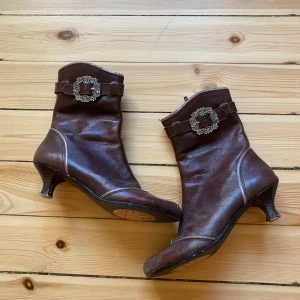 Bruna skinnstövletter med dekorativt spänne - Säljer ett par eleganta bruna skinnstövletter med en liten klack och dekorativa spännen. Perfekta för höst och vinter, dessa stövletter har en klassisk look med en modern twist. De är i bra skick och passar både till vardags och festliga tillfällen.