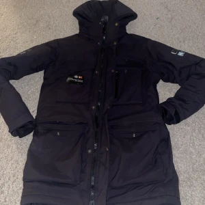 Svart SailRacing Vinter Jacka - Säljer en svart vinterjacka från SailRacing i mycket bra skick. Jackan har en praktisk huva och flera fickor med dragkedja. Perfekt för kalla vinterdagar och håller dig varm och torr. Passar både till vardags och utomhusaktiviteter.