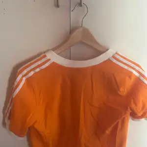 Croppad orange t-shirt från Adidas med vita detaljer. V-ringad hals och de klassiska tre ränderna på ärmarna. Storlek M och har en liten fläck på kragens bak men inte så synlig.