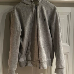 Grå hoodie från Ralph Lauren - Säljer en stilren grå hoodie från Ralph Lauren i storlek M. Den har en dragkedja framtill och en liten broderad logga på bröstet. Perfekt för både höst och vår, med en skön passform och mjukt material. Huvan är justerbar med snören. Passar till alla tillfällen!