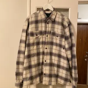 Säljer en snygg rutig overshirt från Casual Friday i storlek XL. Skjortan är i beige och svart med långa ärmar och knappar framtill. Perfekt för höst och vår, och funkar både som jacka och skjorta. Insidan är fodrad för extra värme. Passar perfekt för en avslappnad stil!