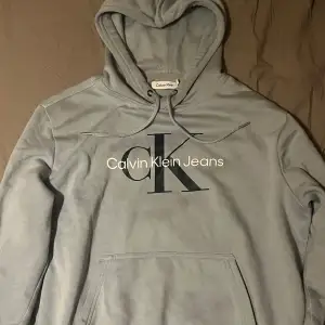 Säljer en snygg grå hoodie från Calvin Klein Jeans. Den har en stor CK-logga på framsidan och en praktisk magficka. Perfekt för en avslappnad stil och passar bra till både jeans och joggers. Huvan har dragsko för justering. Perfekt för kyliga dagar!