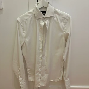 Vit skjorta från Riley - Säljer en stilren vit skjorta från Riley i slim fit. Skjortan har långa ärmar och klassisk krage med knappar framtill. Perfekt för både formella och avslappnade tillfällen. Några små märken finns på framsidan, men annars i bra skick. Passar perfekt till både jeans och kostymbyxor!