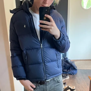 Mörkblå dunjacka från Ralph Lauren - Säljer en snygg mörkblå dunjacka från Ralph Lauren. Jackan har en dragkedja framtill och är långärmad med elastiska muddar. Perfekt för kalla dagar med sin varma vaddering. Den har en klassisk design med en liten logga på bröstet. Passar både till vardags och lite finare tillfällen. Storlek L ungdom passar xs