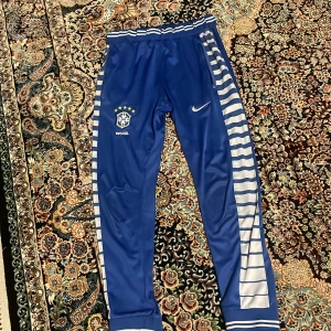 Blå Nike träningsbyxor med Brasilien-logga - Säljer ett par blå träningsbyxor från Nike med Brasilien-logga. Byxorna har vita och blå ränder längs sidorna och en elastisk midja för bekväm passform. Perfekta för fotbollsträning eller avslappnad stil. 🏆