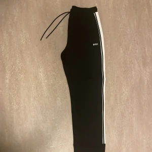Svarta mjukisbyxor från Boss - Snygga svarta mjukisbyxor från Boss med vita sidoremsor och snörning i midjan. Perfekta för en avslappnad stil med en sportig touch. Byxorna har en normal passform lite tight i botten och är tillverkade i ett mjukt material för extra komfort. Storlek M men passar till storlek s