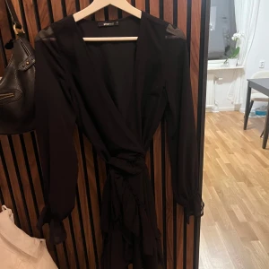 Svart omlottklänning från Gina Tricot - Elegant svart omlottklänning från Gina Tricot med långa ärmar och knytband i midjan. Perfekt för en stilren look. Klänningen har en v-ringad design som ger en sofistikerad touch.