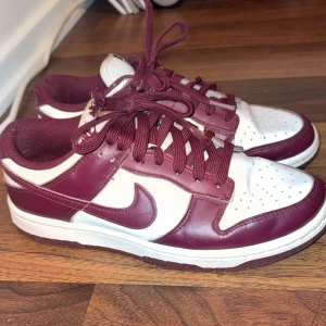 Nike Dunk i vinrött och vitt - Snygga Nike Dunk sneakers i vinrött och vitt. Skorna har en klassisk design med snörning och det ikoniska Nike-märket på sidan. Perfekta för en stilren look.