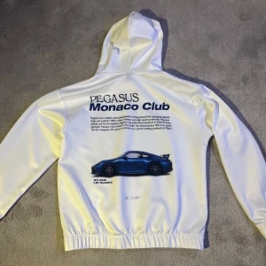 Vit hoodie med tryck från Pegasus Monaco Club - Säljer en vit hoodie från Pegasus Monaco Club med tryck av en Porsche och text på ryggen. Framsidan har en liten logga i blått. Hoodien har en klassisk design med ett snyggt tryck. Tröjan är oanvänd 
