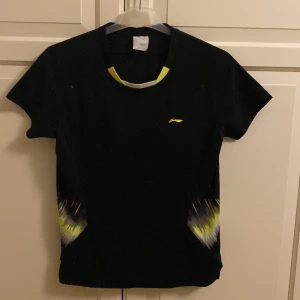 Svart kortärmad t-shirt med mönster från Li-Ning - Snygg svart t-shirt med korta ärmar och coola mönster i gult och vitt på sidorna. Perfekt för en sportig look med en touch av stil. Den har en rund halsringning med gul detalj. Från Li-Ning. I fint skick. Storlekslappen är borttvättad, men passar som XS/S. 