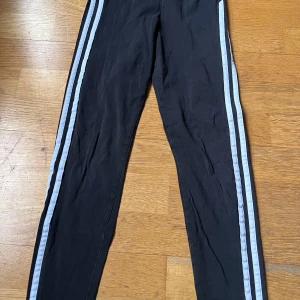 Svarta leggings med ränder från Adidas - Snygga svarta leggings från Adidas med vita ränder längs sidorna. Perfekta för träning eller en avslappnad dag. De har en tight passform och är tillverkade i ett bekvämt syntetmaterial. (För barn, 11-12 år)