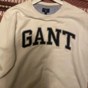 Vit sweatshirt från GANT - Säljer en stilren vit sweatshirt från GANT med deras logga i svart på bröstet. Tröjan har en klassisk rund hals och långa ärmar. Perfekt för en avslappnad stil. Inga skador alls, bara vilar o garderoben! 