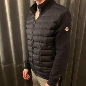 Marinblå cardigan från Moncler - Snygg cardigan från Moncler med dragkedja framtill. Jackan har långa ärmar och en quiltad design som ger en stilren look. Perfekt för kyligare dagar. Jackan har en liten defekt då sömmen nere vid höger handled har lossnat, inget som syns när man använder den, går självklart att sy ihop lätt