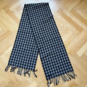 Rutig halsduk från A. Christensen - Snygg rutig halsduk i svart och grått från A. Christensen. Den har fransar i ändarna och ett klassiskt mönster som passar perfekt för kyliga dagar. Perfekt accessoar för att hålla stilen och värmen.