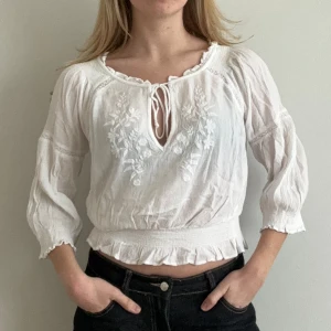 Vit blus med broderi - Charmig vit blus med broderade detaljer och knytning framtill. Blusen har trekvartsärmar och en elastisk midja som ger en fin passform. Perfekt för en avslappnad och stilren look.