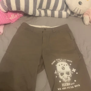 Bruna shorts med tryck från Zeero One - (32tum är lite som ett M)Coola bruna shorts från Zeero One med punkinspirerat tryck av en dödskalle och text. Shortsen har praktiska fickor och en dragkedja framtill. Thriftad själv och bara några lösa trådar men i perfekt skick, känns lite mindre än 32 tum.