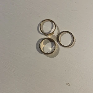 Tre guldiga ringar - Säljer tre stilrena guldiga ringar. En av ringarna har en slät yta med en liten platta, den andra har ett flätat mönster och den tredje är tunn med en vågig kant. Perfekta för att bära tillsammans eller var för sig för en elegant look. 70 kr för alla tre 