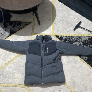 Grå dunjacka från Sail Racing - Säljer en stilren grå dunjacka från Sail Racing. Jackan har en dragkedja framtill och är långärmad. Perfekt för kyligare dagar med sin quiltade design. Den är tillverkad i syntetmaterial och har en modern look.