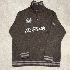 St.Moritz Halvzip  - Säljer en brun stickad tröja med St. Moritz tryck med dragkedja och broderad logga på bröstet. Tröjan har långa ärmar och ribbade muddar i grått vid ärmslut och nederkant. Perfekt för kyliga dagar! Vid minsta fråga eller fundering hör av er🤝