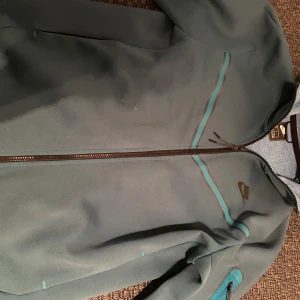 Grön jacka från Nike - Säljer en grön jacka från Nike med dragkedja och stilrena detaljer i turkos. Jackan har en normal passform och är perfekt för vardagsbruk. Den har långa ärmar och en diskret Nike-logga på bröstet.