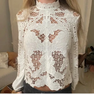 Vit spetsblus från Zara - Säljer en elegant vit spetsblus med hög krage och långa ärmar. Blusen har ett vackert blommigt mönster och är genombruten vilket ger en stilren look. Perfekt för att lyfta vilken outfit som helst.