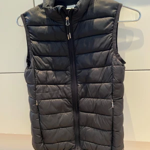 Svart väst från CRW stl 158/164 - Säljer en svart ridväst från CRW med dragkedja framtill. Västen har en quiltad design och är perfekt för kyligare dagar. Den är stilren och lätt att matcha med olika outfits. Passar både för vardag och utomhusaktiviteter.